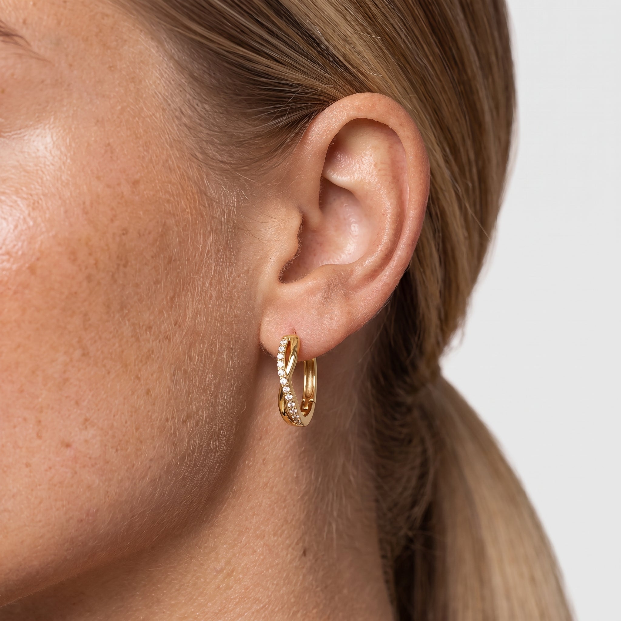 Gold-tone twisted crystal huggie hoop earring worn in the ear